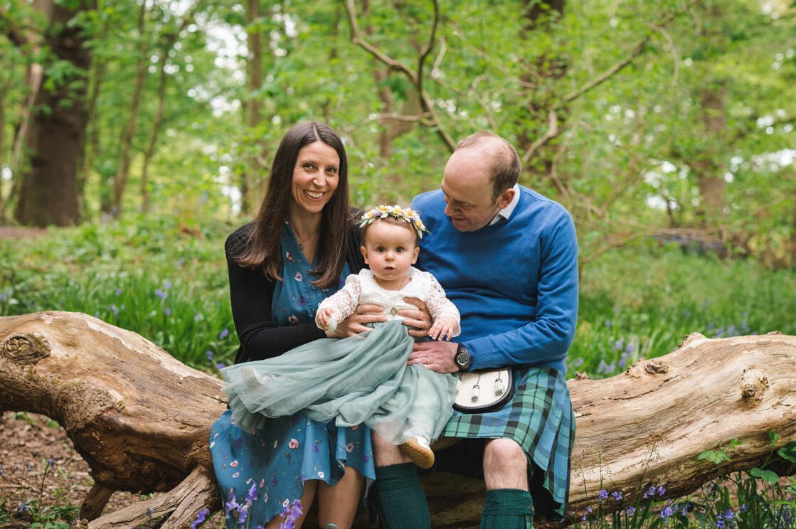 Photo of my family from my daughter's naming ceremony. I'm the woman on the left with long dark hair and smiling, wearing a blue dress with flowers. Husband is on the right and wearing a blue jumper with a kilt and sporran. Our daughter is between us.