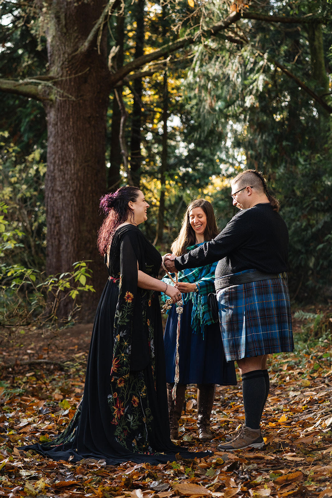 I'm standing in front of a couple in a woodland vow renewal ceremony as a tie their handfasting cord. We are all smiling and laughing. There are autumn leaves all around us on the forest floor and beautiful sunlight streaming in from the left.