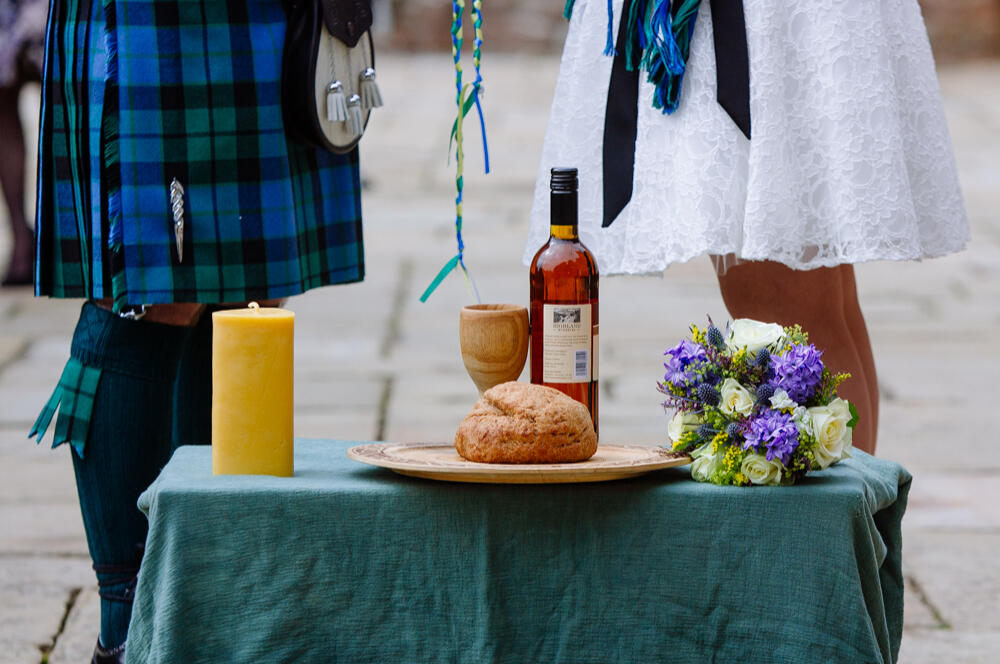 A couple stand behind a low altar table holding a candle, wooden plate with a round loaf, wooden goblet, bottle of mead, and a wedding bouquet. One of the couple wears a blue and green kilt and the other wears a knee-length white wedding dress. A green and